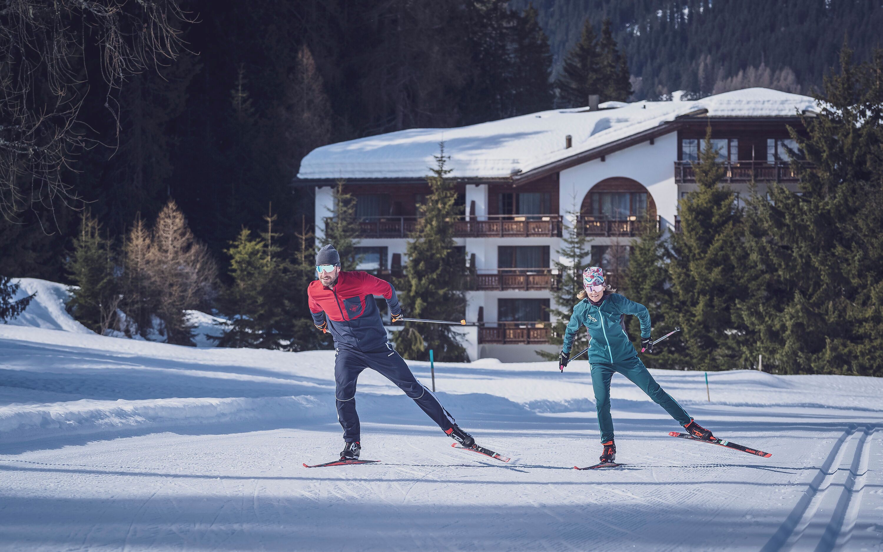 Two skiers on a snowy slope with a building in the background.