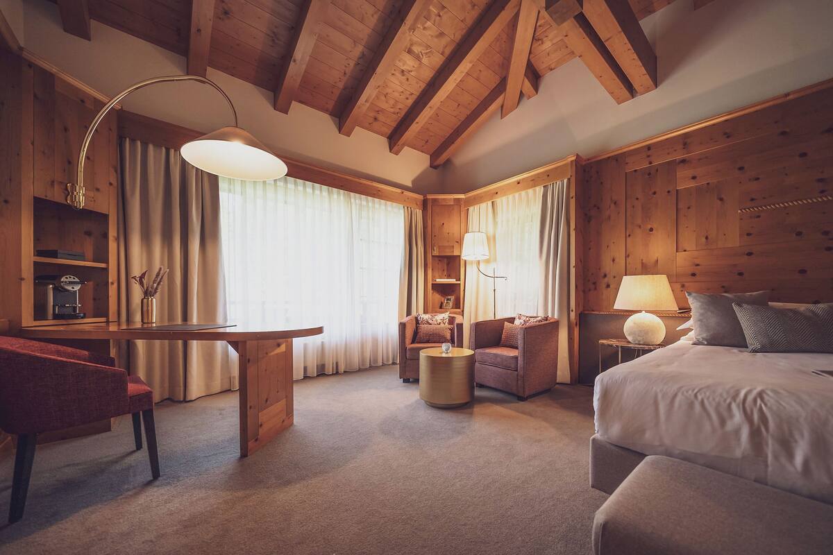 A hotel room with a wooden ceiling, a bed, a desk, two armchairs, a small table, and a lamp.
