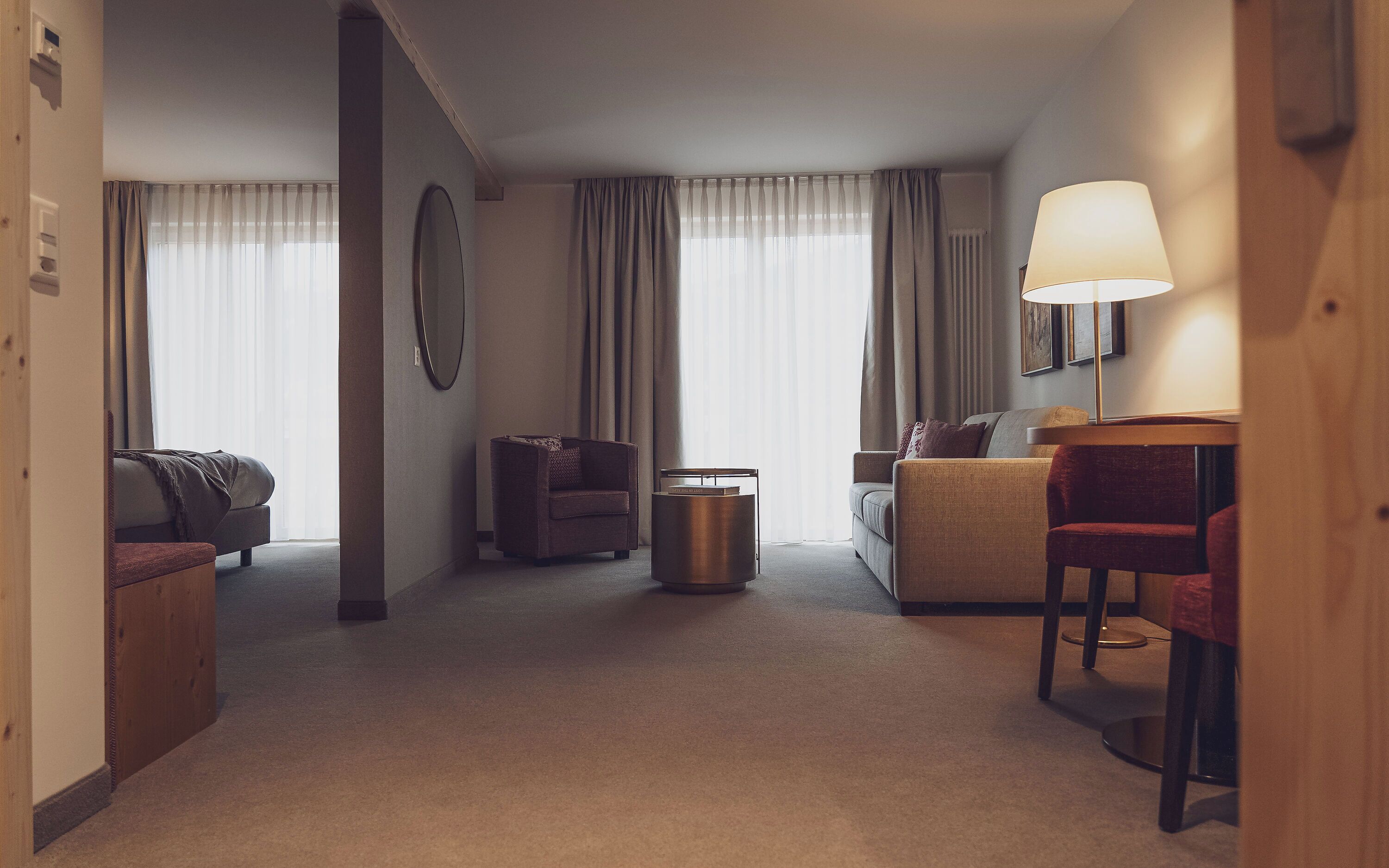 A hotel room with a sofa, armchair, and a small table with a lamp.