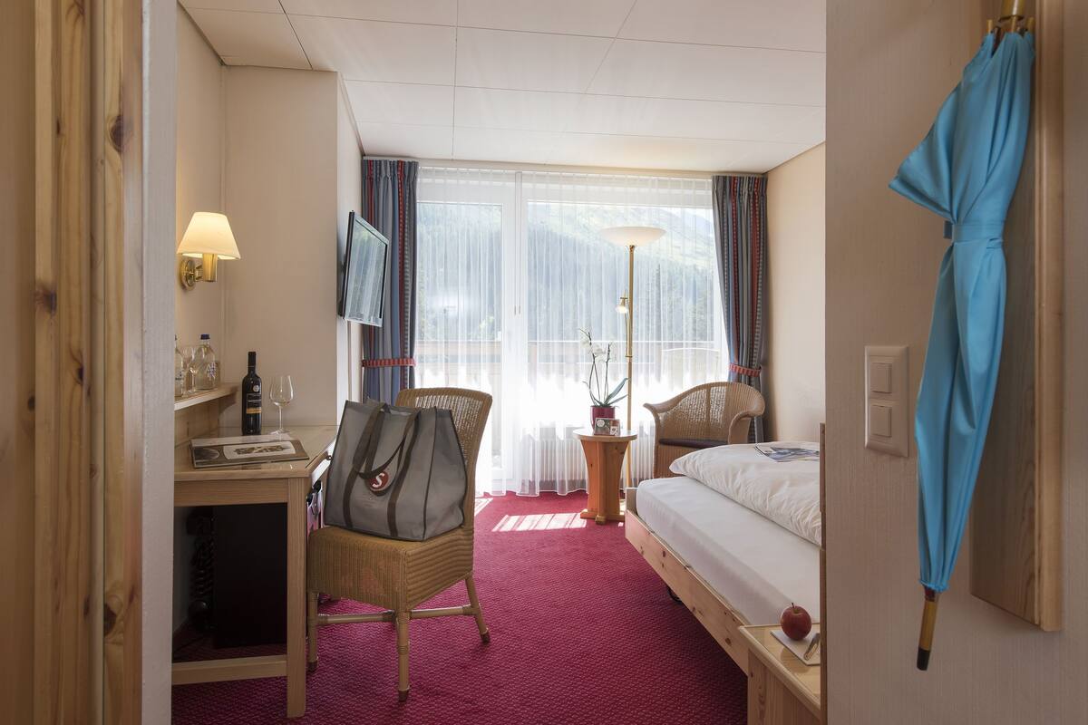 A hotel room with a bed, a desk with a lamp, a chair, a small table with a bottle and glasses, and a window with curtains.