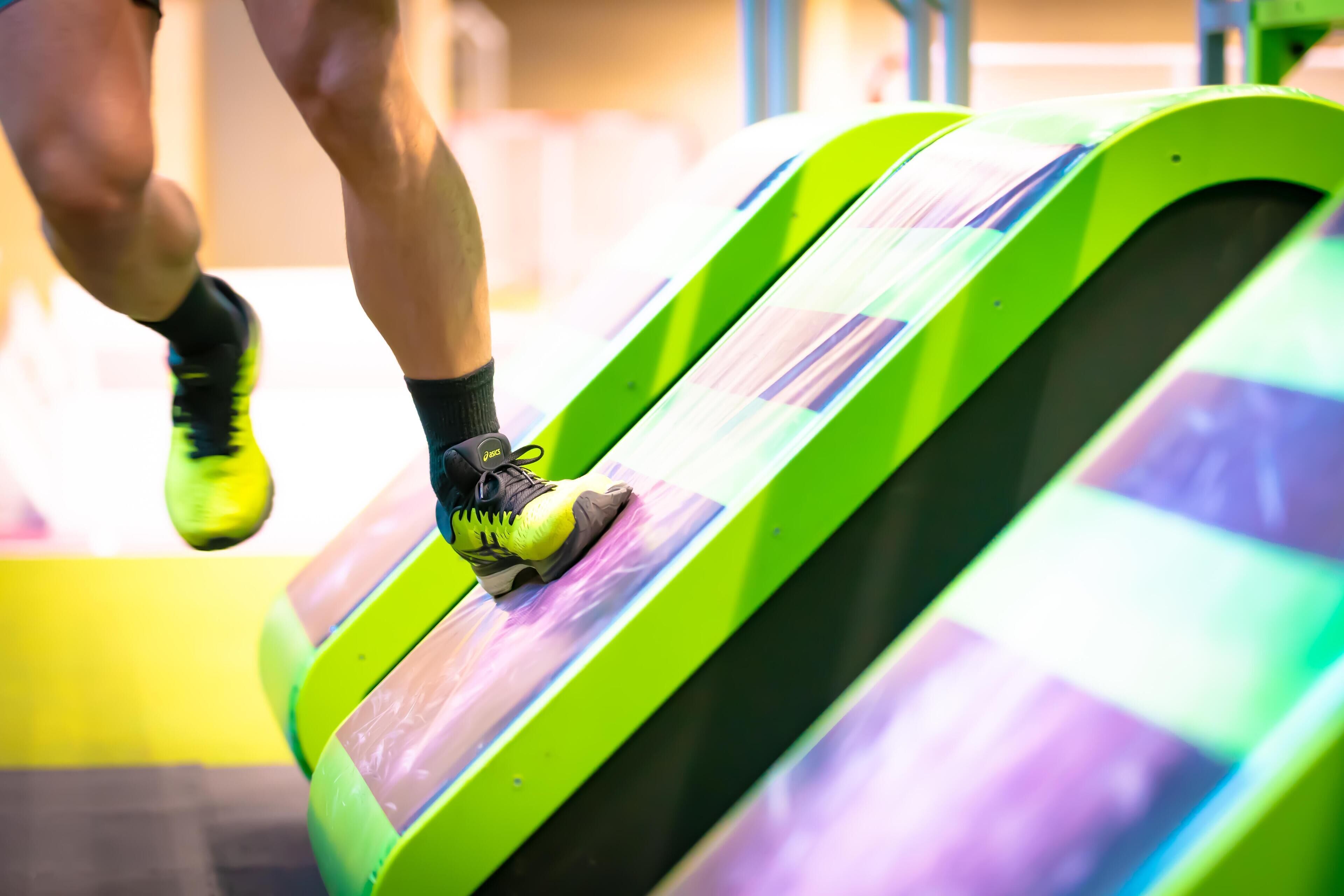 Une personne court sur un tapis de course vert et incurvé, avec un fond rayé de violet et de blanc.