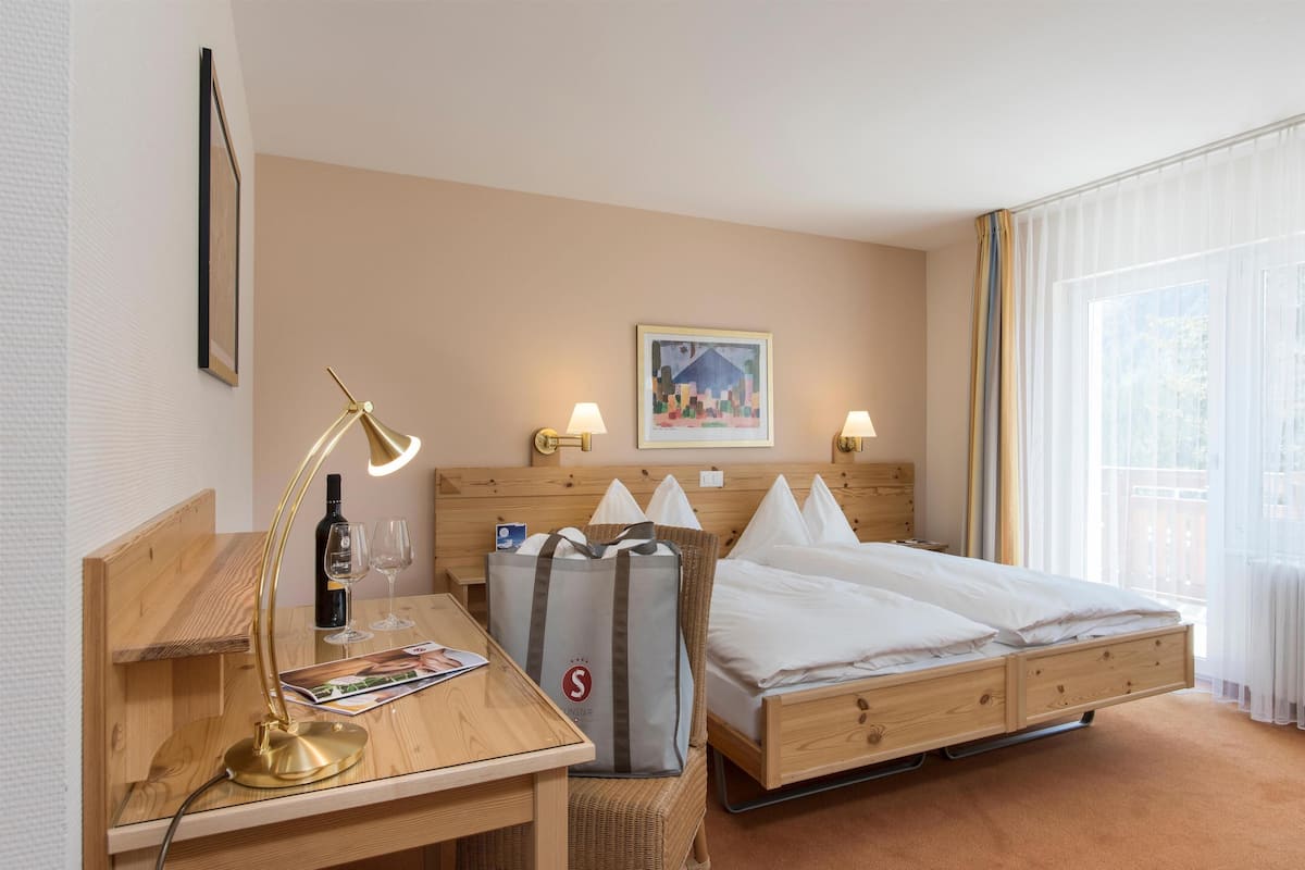 A hotel room with a wooden desk, a bed with white bedding, a headboard with wall-mounted lamps, a framed picture on the wall, and a large window with sheer curtains.