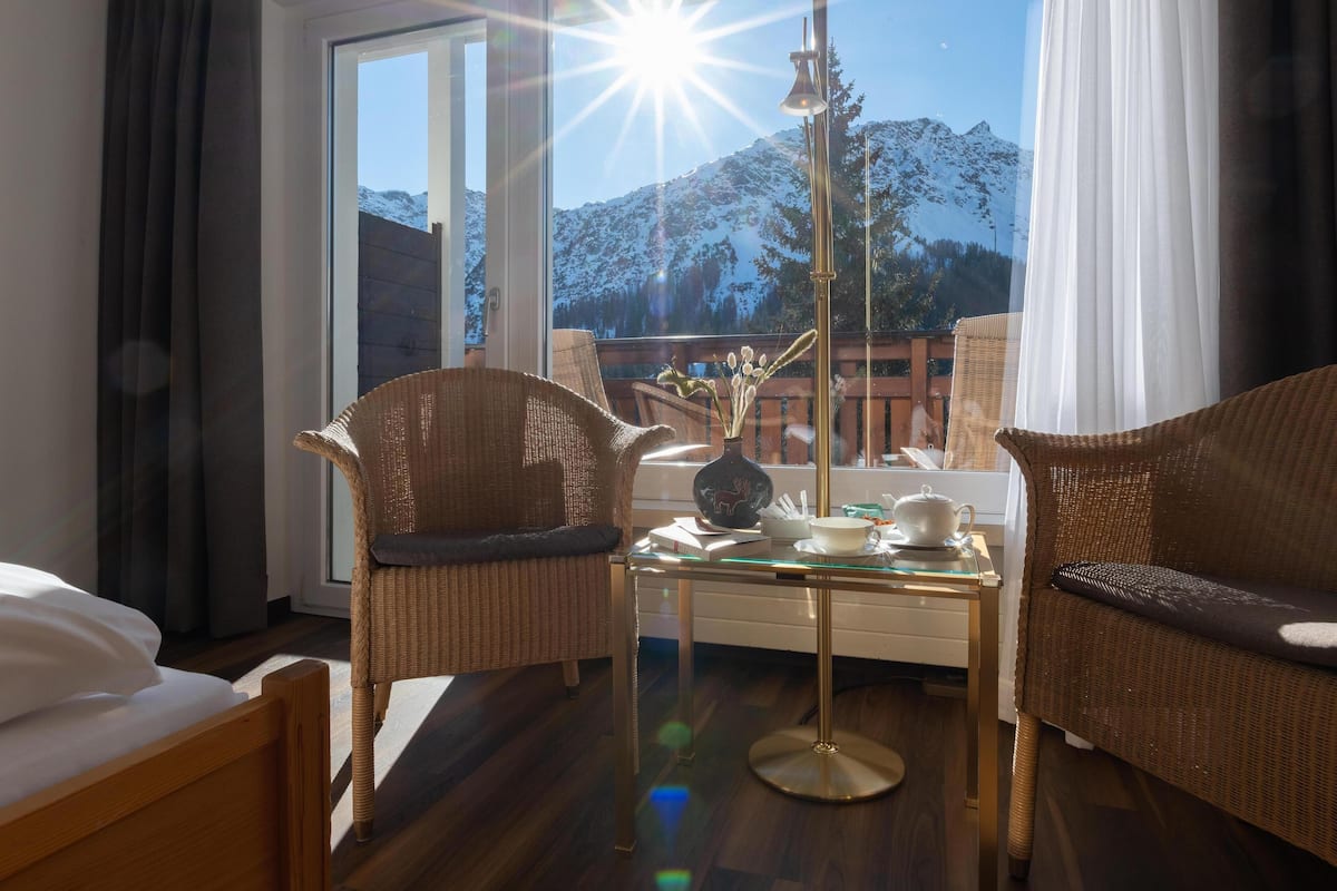 A hotel room with a bed, two wicker chairs, a glass coffee table, and a view of snow-capped mountains.