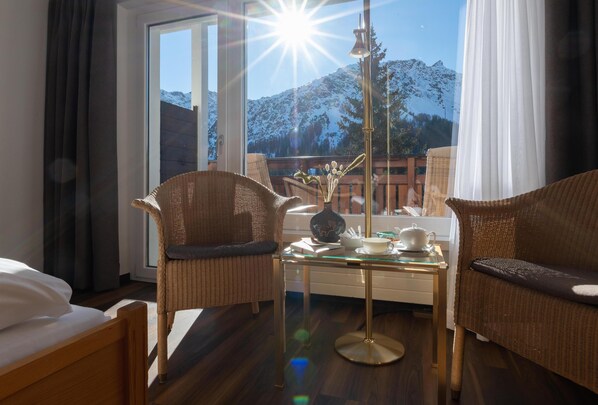 A hotel room with a bed, two wicker chairs, a glass coffee table, and a view of snow-capped mountains. - Sunstar Hotel Arosa (Arosa)