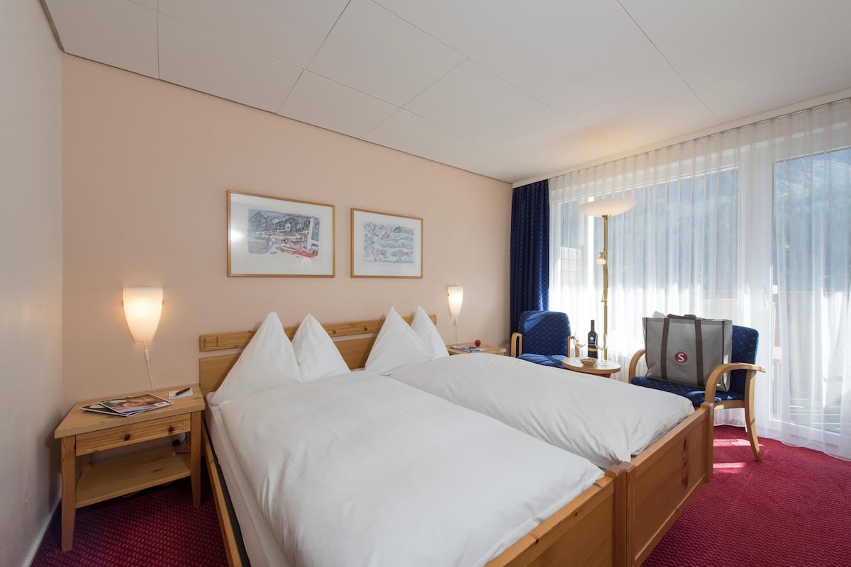 A hotel room with two beds, a wooden nightstand, a chair, a table, and a large window with sheer curtains.
