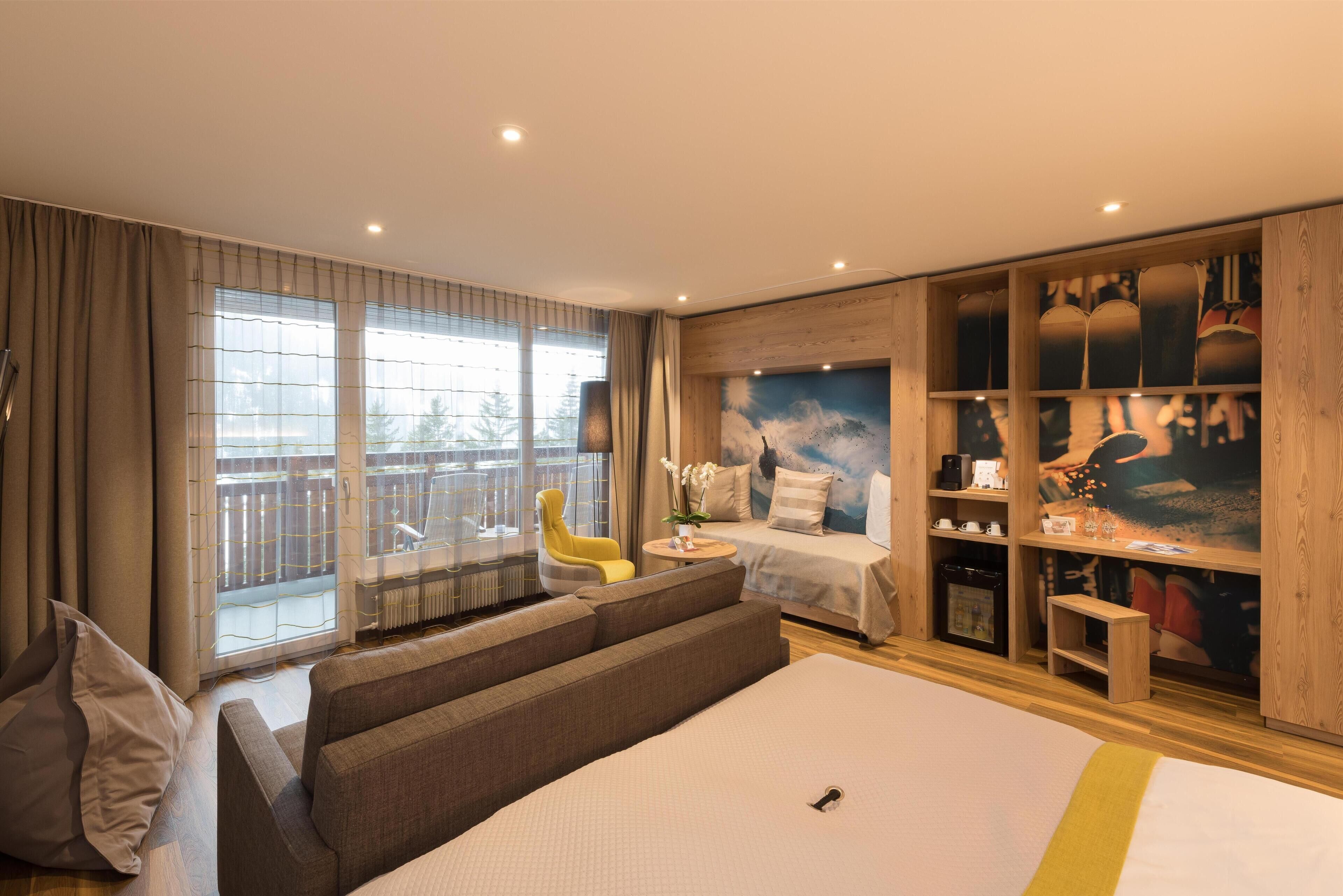 A modern hotel room with a large bed, a sofa, a small table, and a chair. There is a window with curtains and a view of the outside.