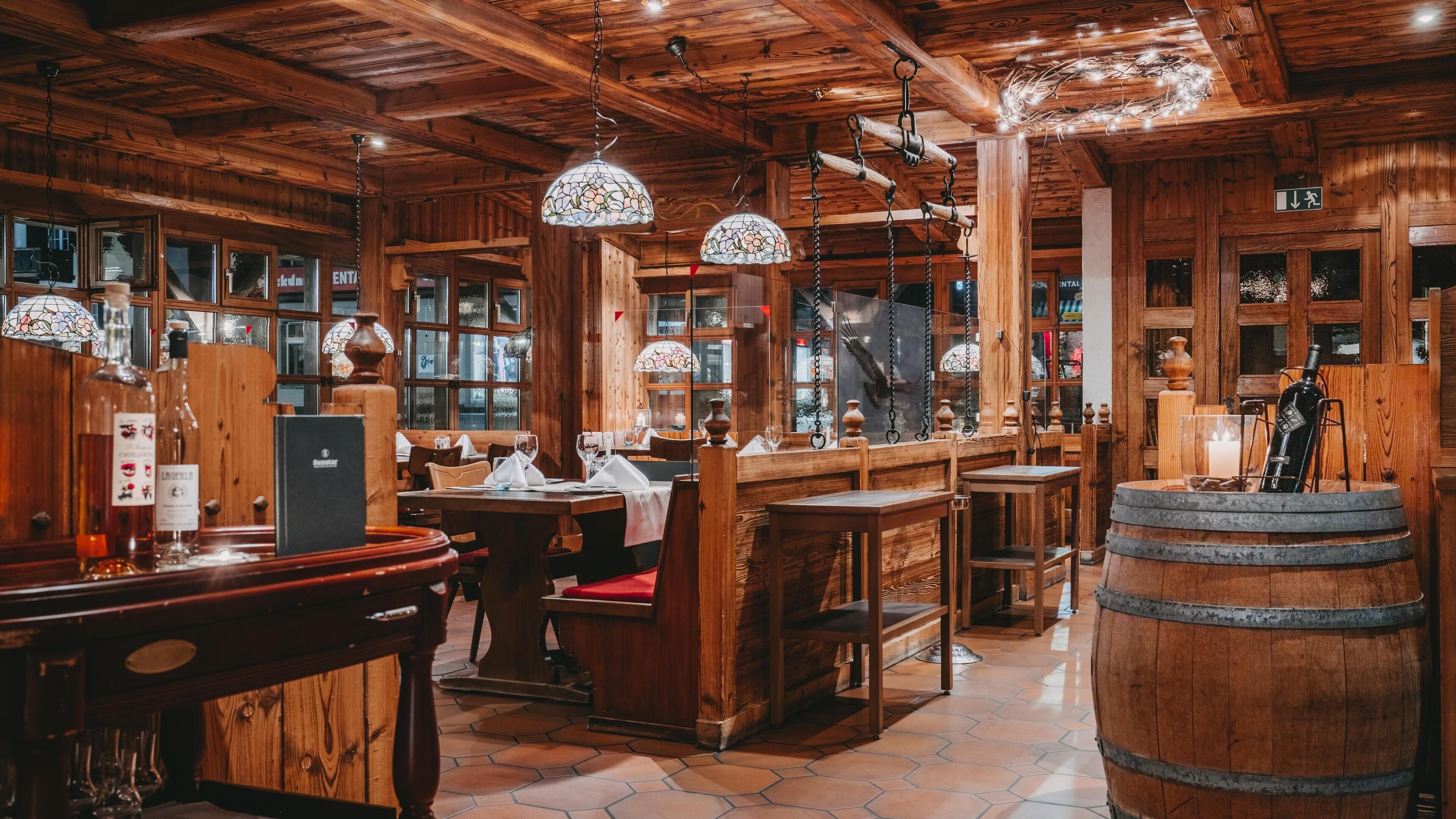 A rustic wooden interior with wooden tables and benches, hanging light fixtures, and a large barrel.