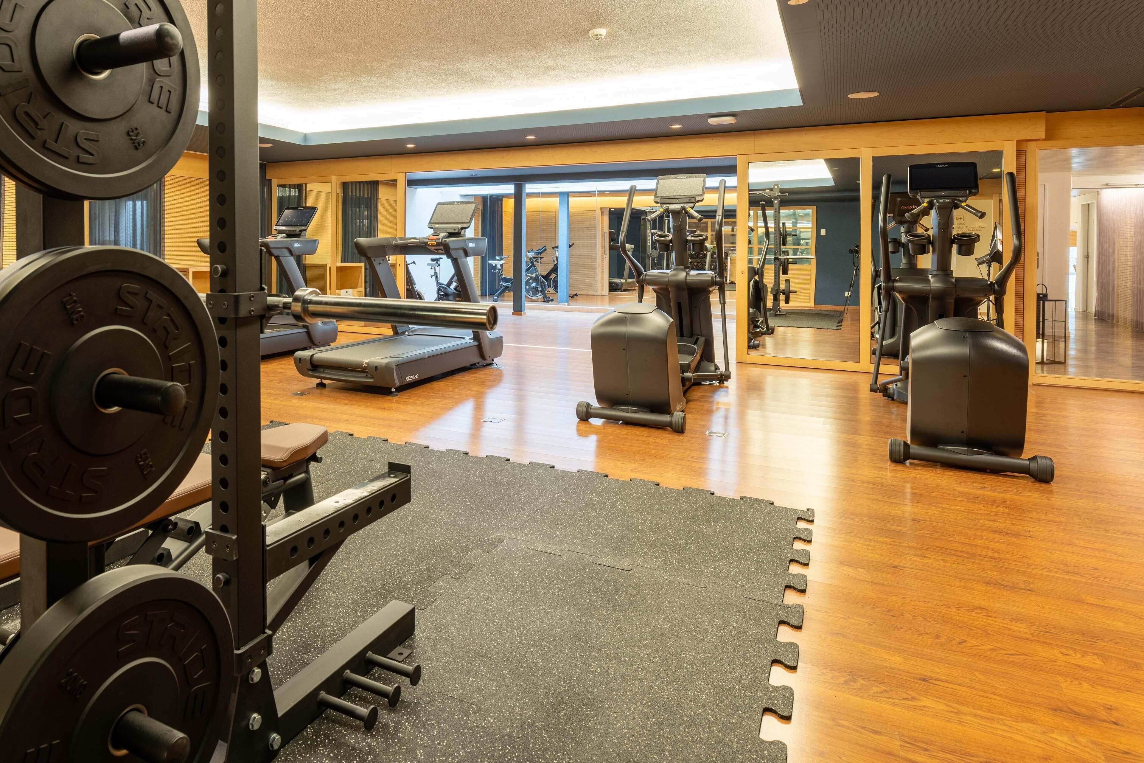 Ein Fitnessstudio mit unterschiedlichen Trainingsgeräten, darunter Laufbänder und Kraftgeräte, sowie einem Holzboden.