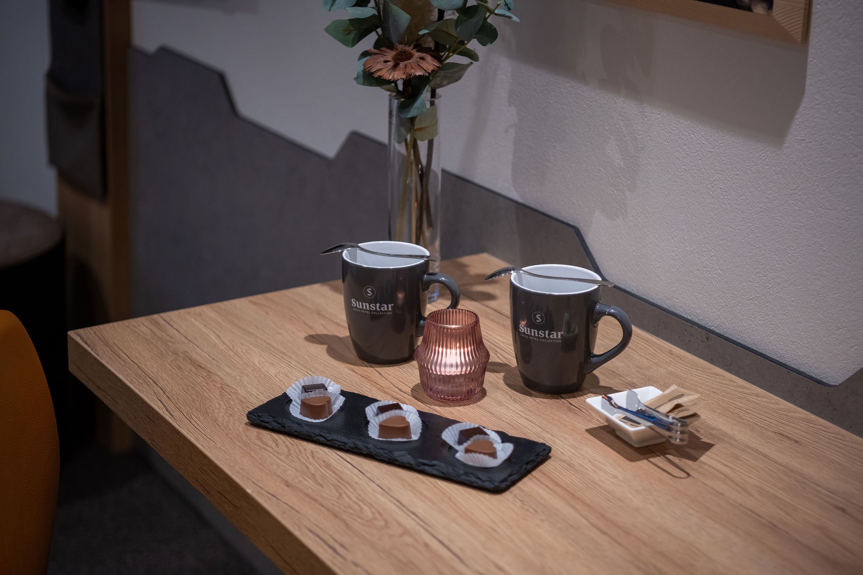 Una mesa de madera con dos tazas de café, un florero con una flor y una bandeja con bombones.