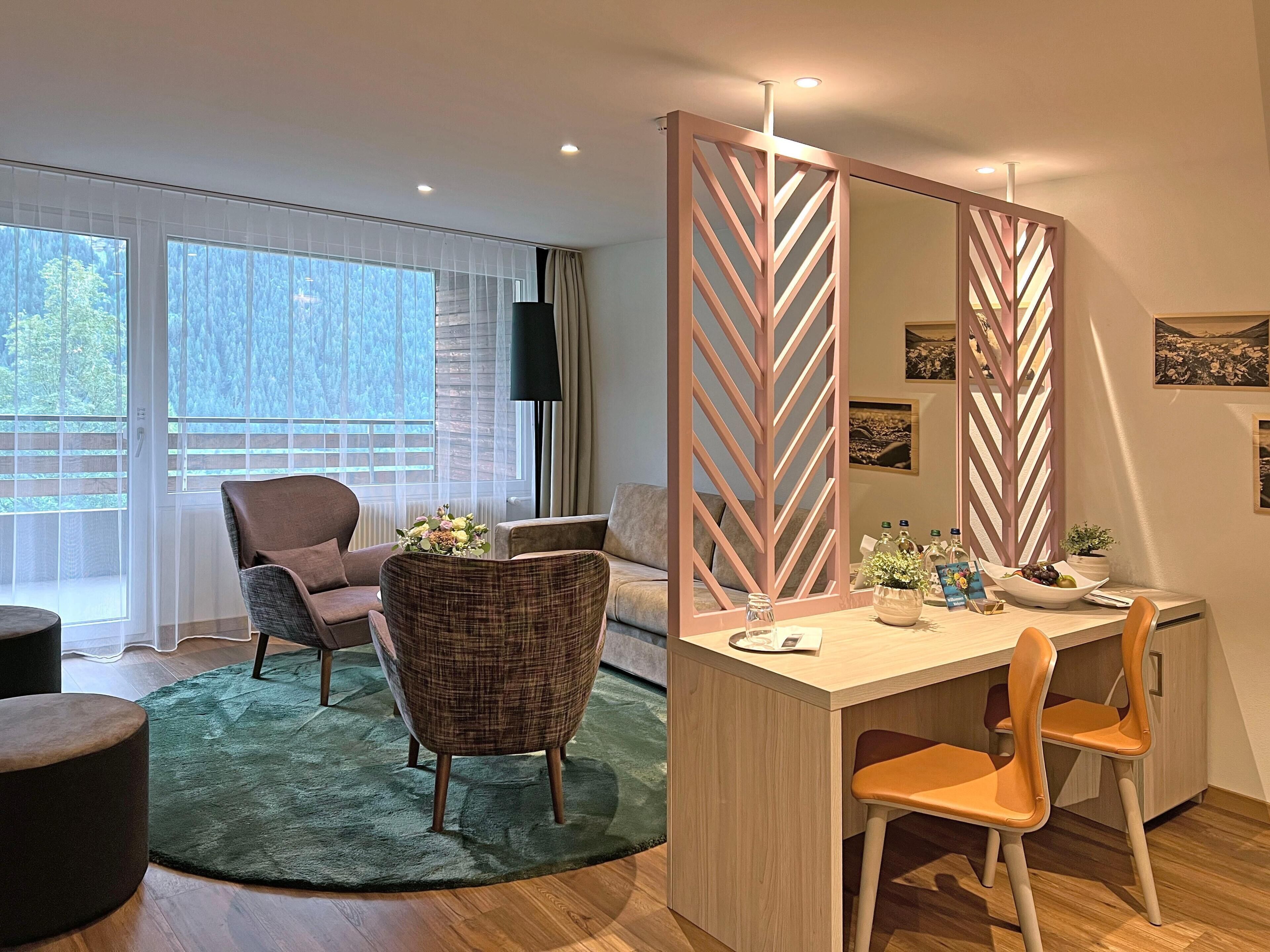 Habitación de hotel moderna con un escritorio de madera, dos sillas, un espejo grande y vistas al exterior a través de amplios ventanales.