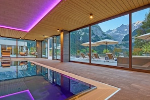 Una piscina interior moderna con paredes de vidrio transparente, suelo de madera y vistas a montañas.