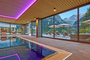Piscina interior moderna com paredes de vidro transparente, piso de madeira e vista para montanhas.