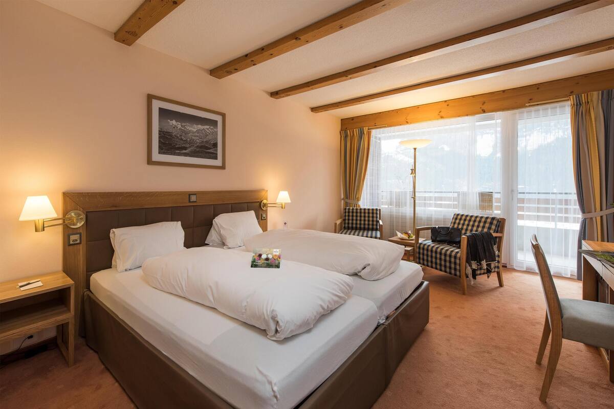 Une chambre d’hôtel avec un grand lit, deux lampes de chevet, une chaise, un bureau et une fenêtre avec des rideaux.
