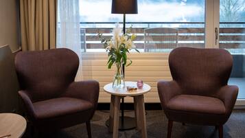 Une pièce avec deux fauteuils marron, une petite table ronde ornée d’un vase de fleurs, une lampe et une vue sur un balcon avec une balustrade.