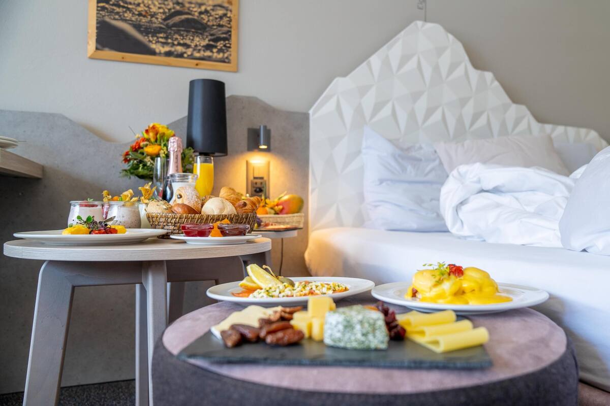 Une chambre d’hôtel avec un lit soigneusement fait, une table à manger avec des aliments pour le petit-déjeuner, et une petite table ronde avec du fromage et de la charcuterie.