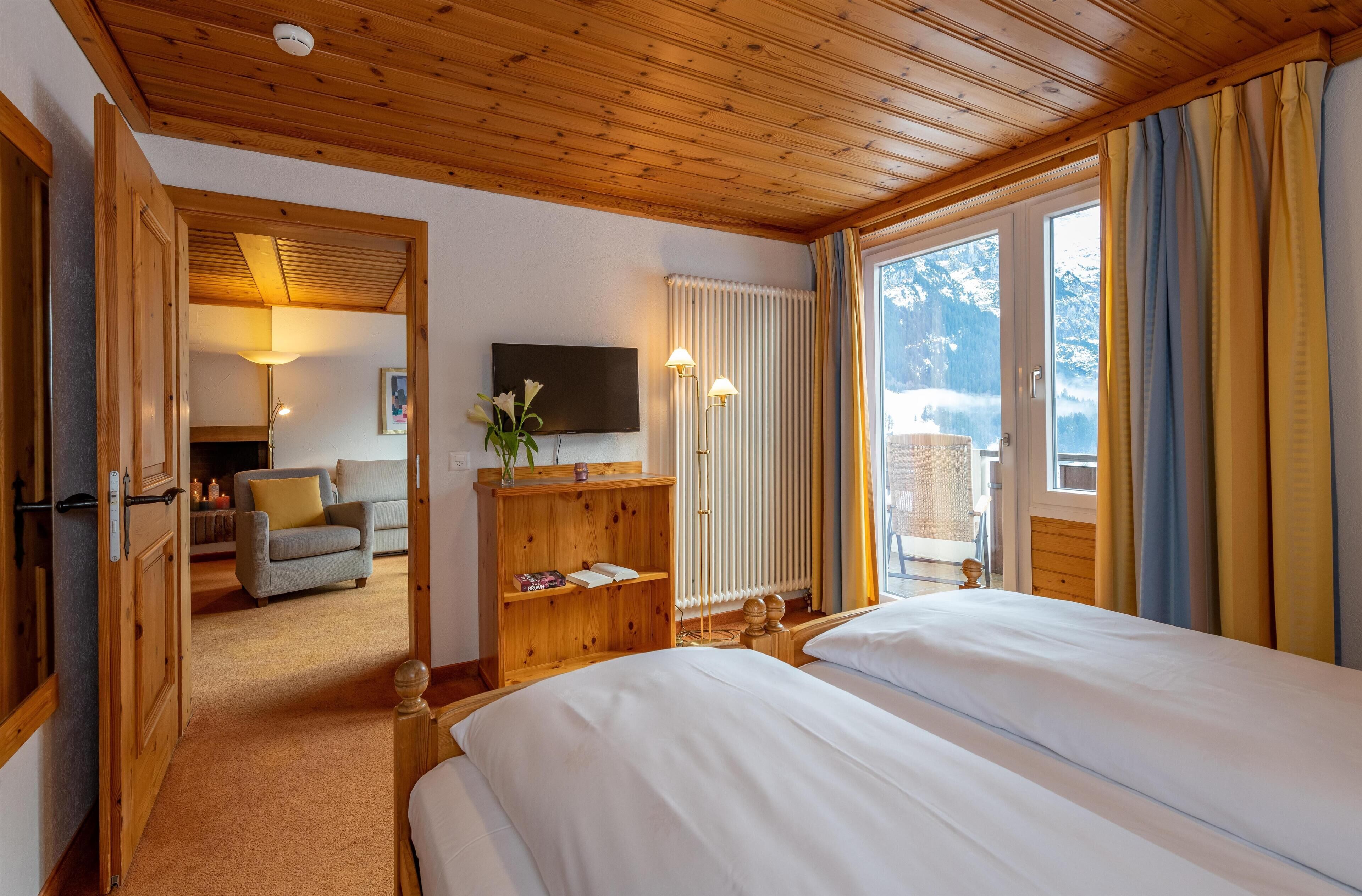 A hotel room with wooden ceilings, a bed, a TV, a radiator, and a view of snow-capped mountains.