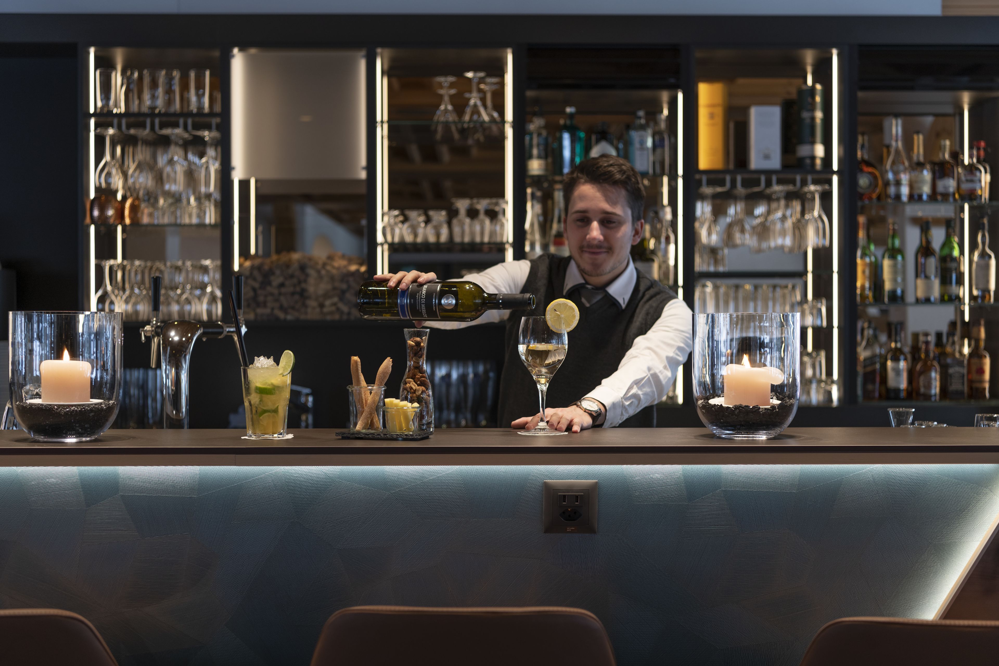 A bar area with a bartender pouring a drink, glasses, and bottles on shelves.