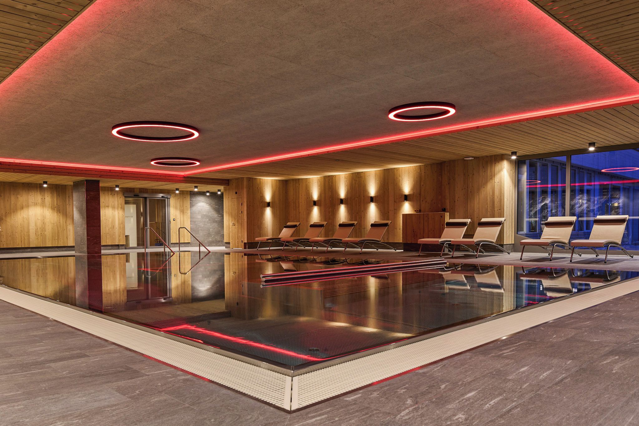 Una piscina interna moderna con rivestimenti in legno, illuminazione con luci rosse e poltrone relax.