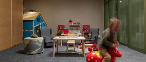 Una sala de juegos con una casita azul, una mesa, sillas y un niño montando un caballo de juguete rojo.