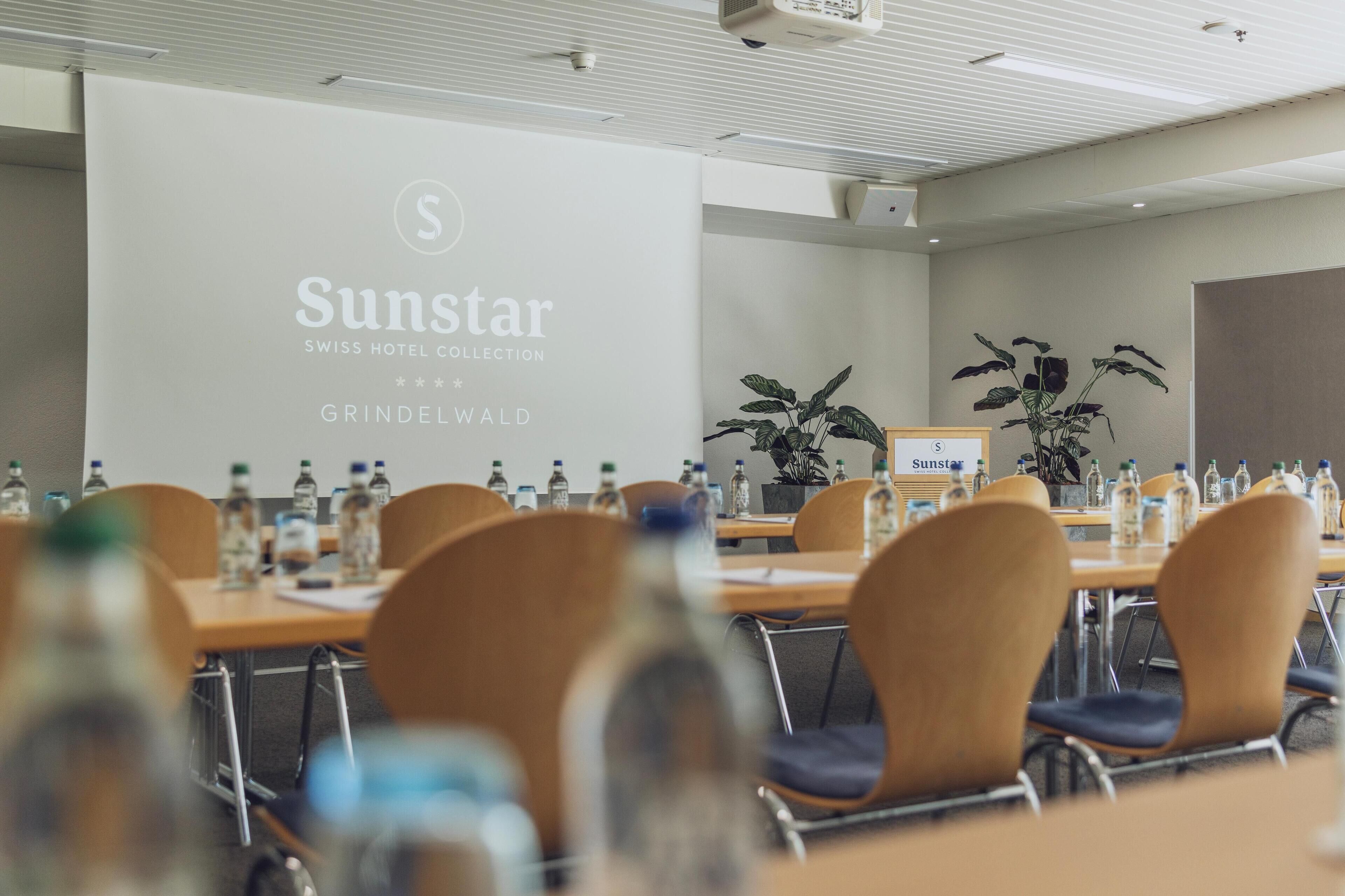 A conference room with tables, chairs, and water bottles. A large screen displays the Sunstar logo and name.