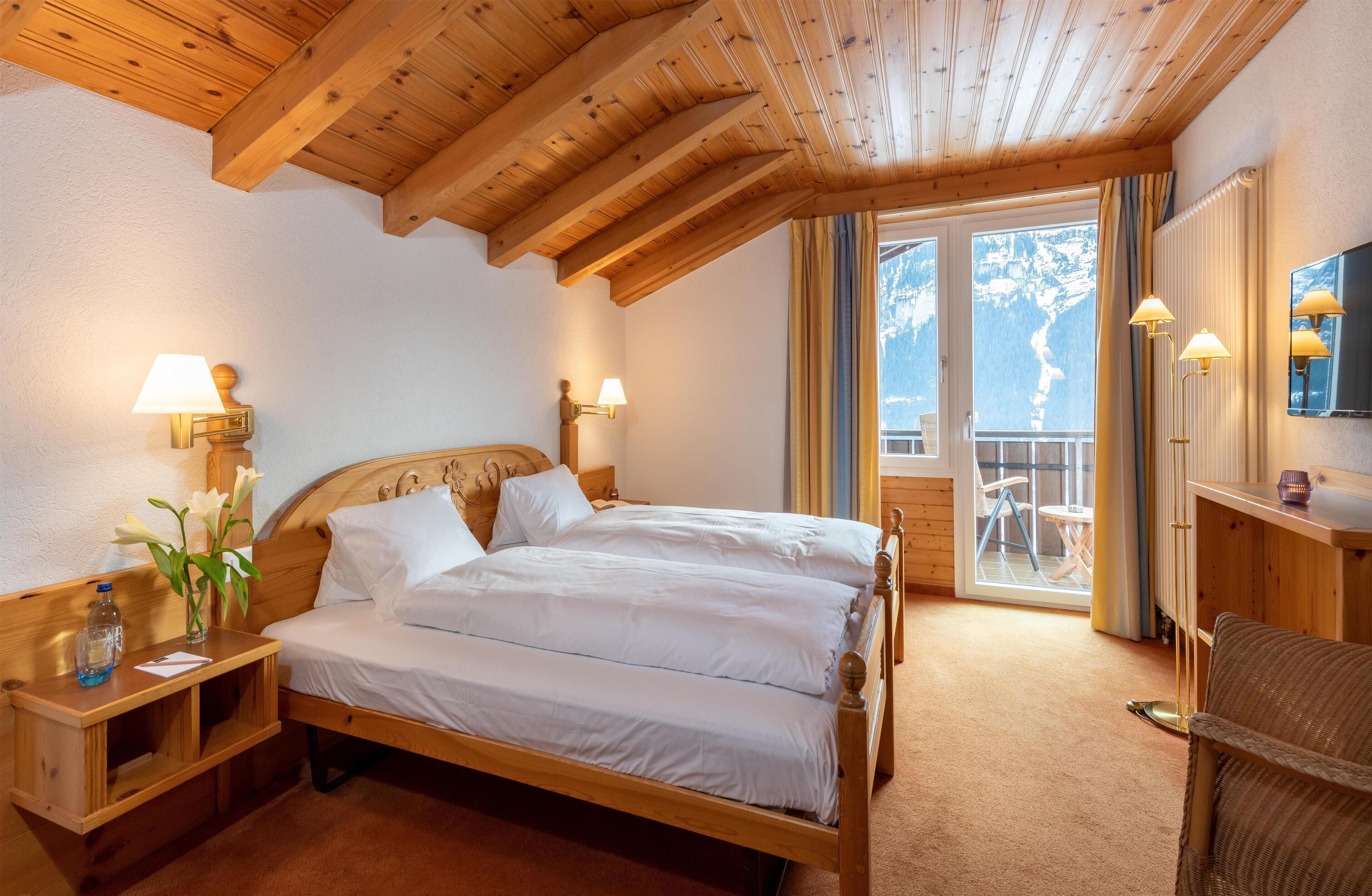Ein schlicht eingerichtetes Schlafzimmer mit Holzbett, Nachttischlampen, einem kleinen Tisch mit Blumenvase und Blick auf einen Balkon mit Stuhl.