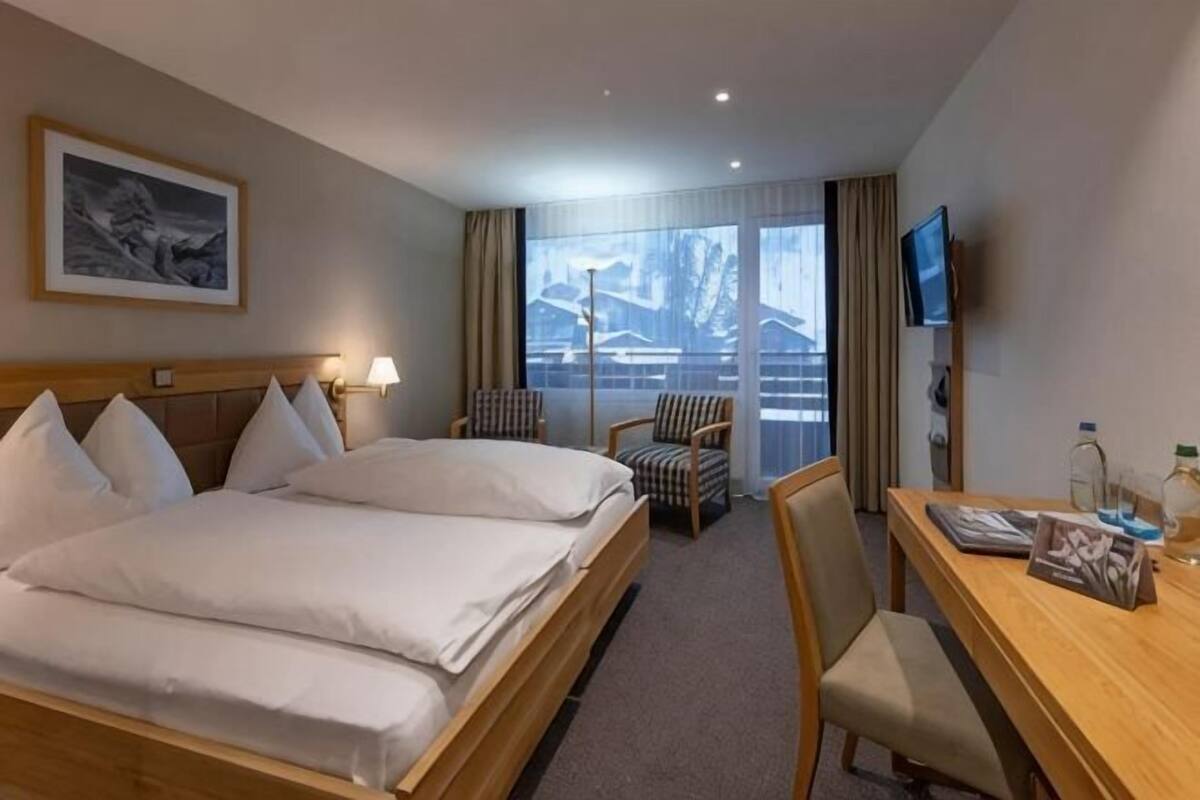 Une chambre d’hôtel avec un grand lit, un bureau, une chaise, une télévision et une fenêtre donnant sur un paysage enneigé.