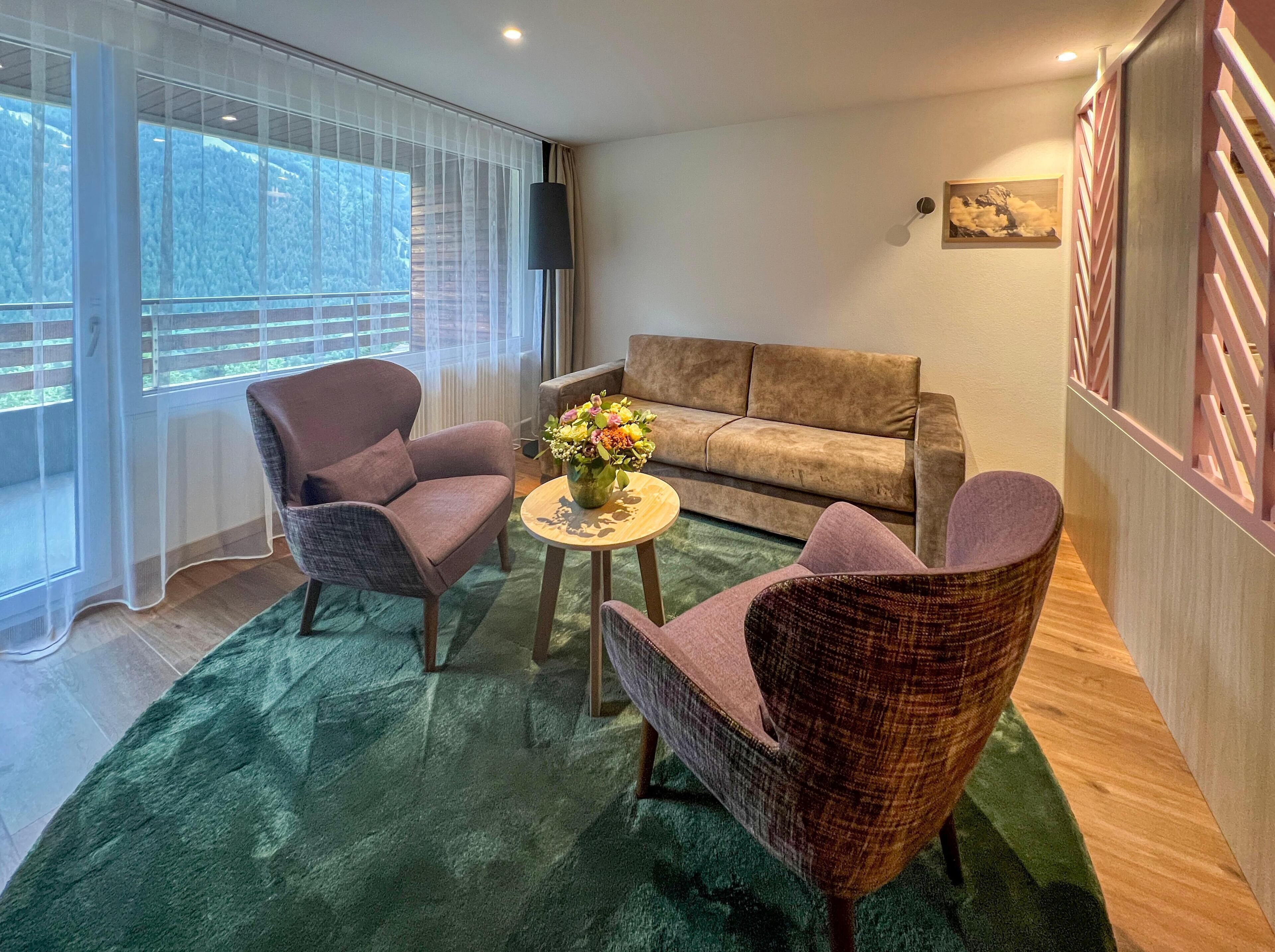 Ein Wohnzimmer mit grünem Teppich, zwei Sesseln, einem Sofa und einem kleinen runden Tisch mit Blumenvase.