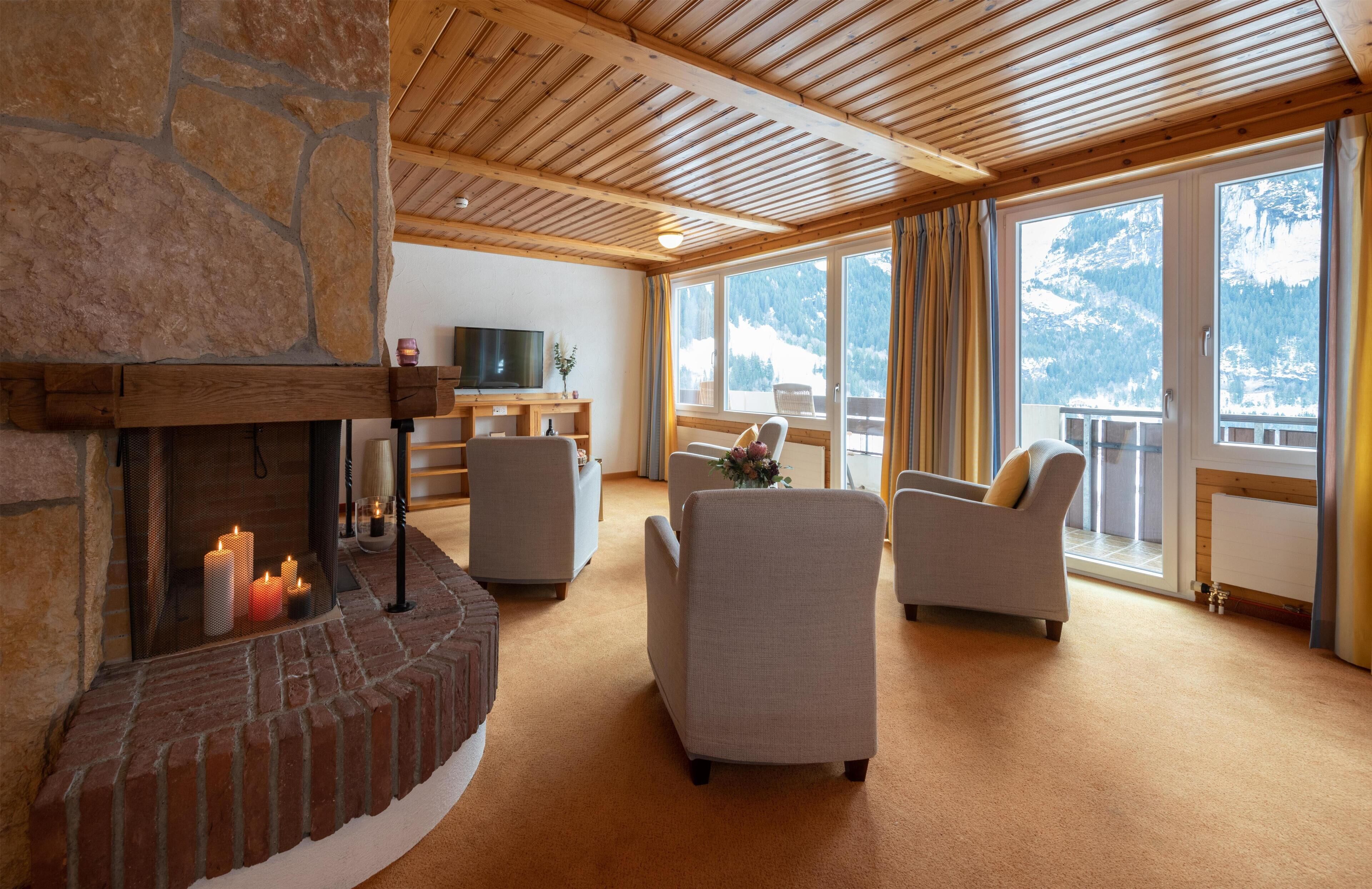 Ein gemütliches Wohnzimmer mit Kamin aus Stein, bequemen Sitzmöglichkeiten, einem Fernseher und großen Fenstern mit Blick auf die Berge.