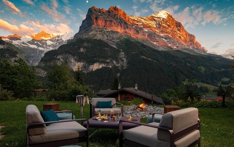 A cozy outdoor seating area with a fireplace, surrounded by a mountainous landscape and a clear sky.