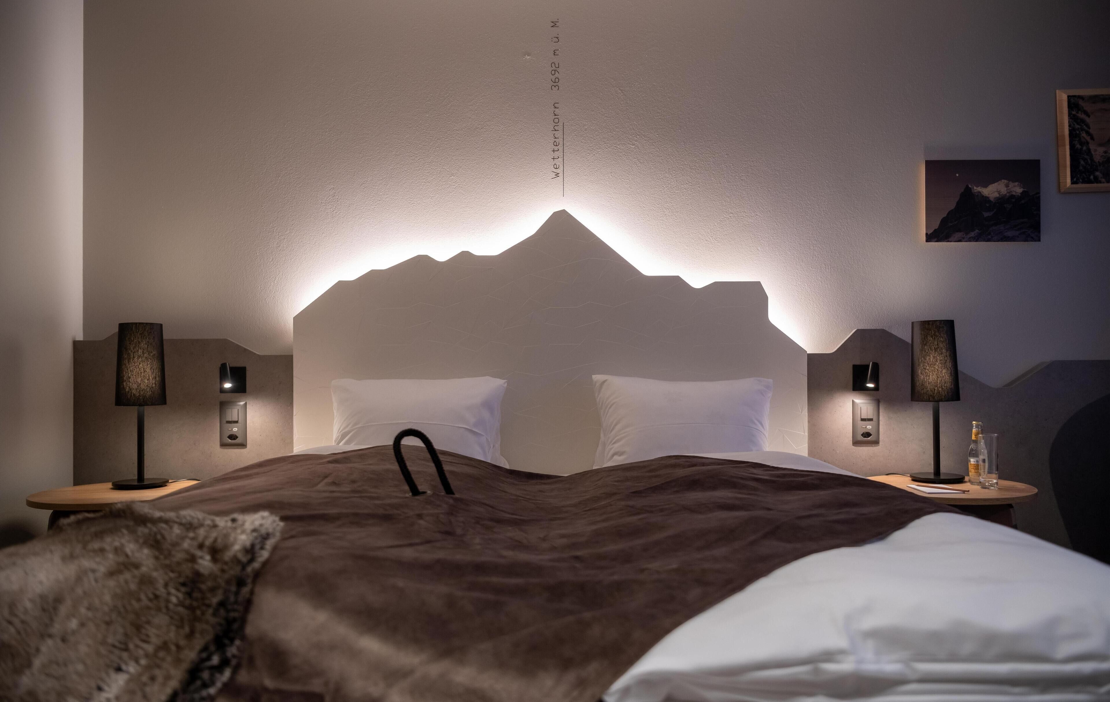 A modern hotel room with a mountain-themed headboard, two bedside tables with lamps, a fluffy brown throw blanket, and a framed picture on the wall.