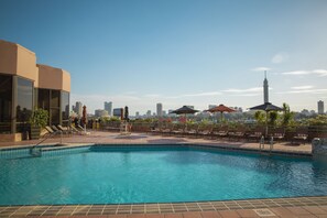 A rooftop pool with lounge chairs and umbrellas, overlooking a city skyline.