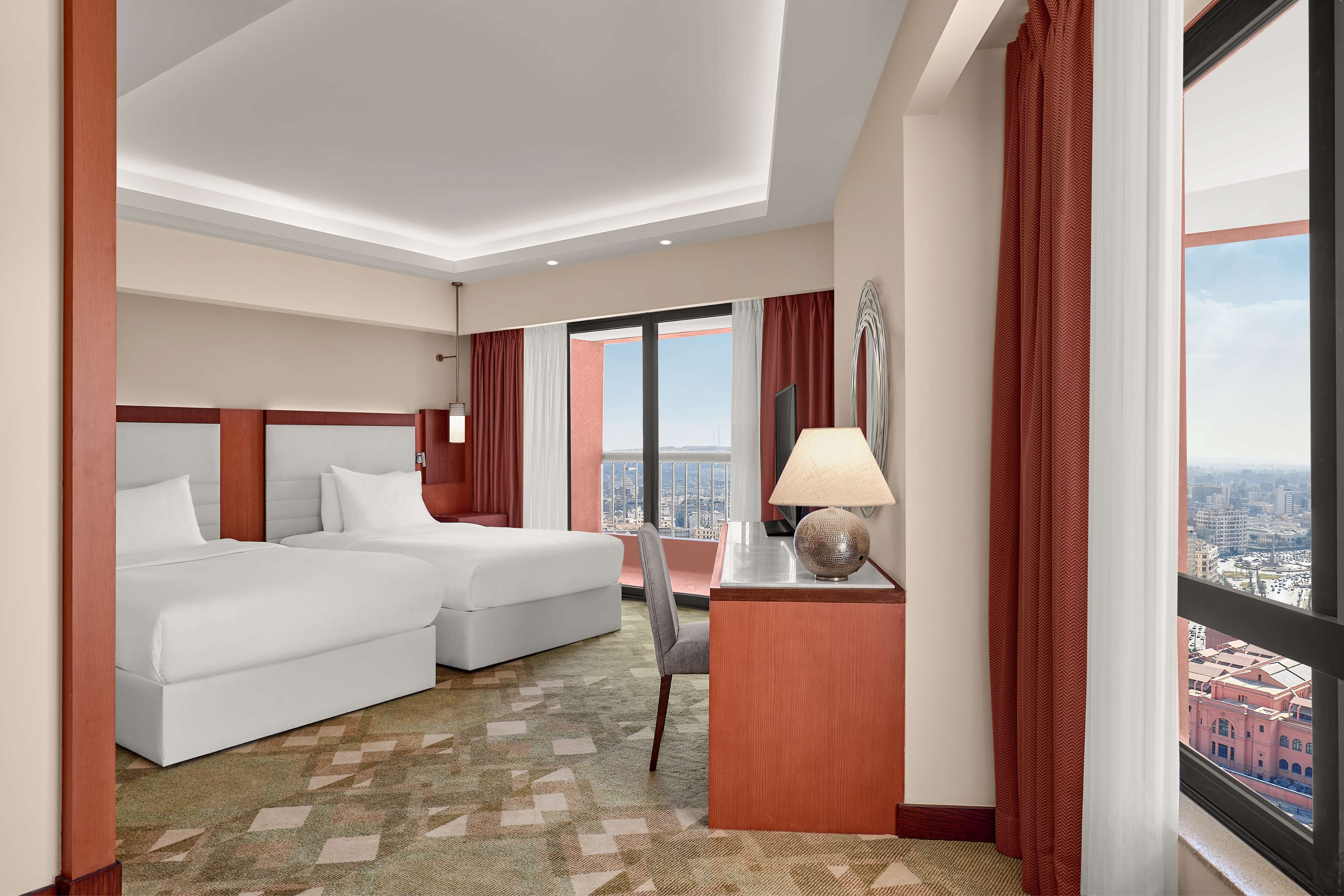 A hotel room with two beds, a desk with a lamp, a chair, and a large window offering a city view.