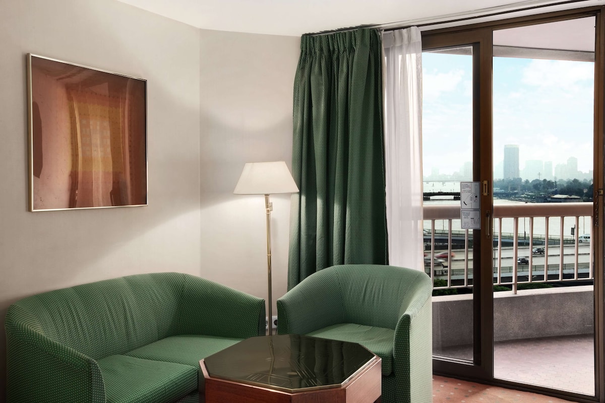A hotel room with a green sofa, a small table, a lamp, and a view of the city through a balcony.