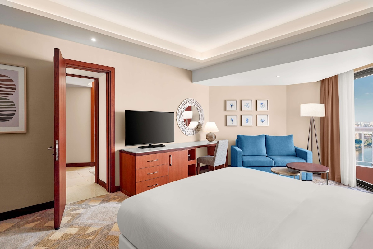 A hotel room with a large bed, a TV, a desk with a chair, a blue sofa, and a view of the city.