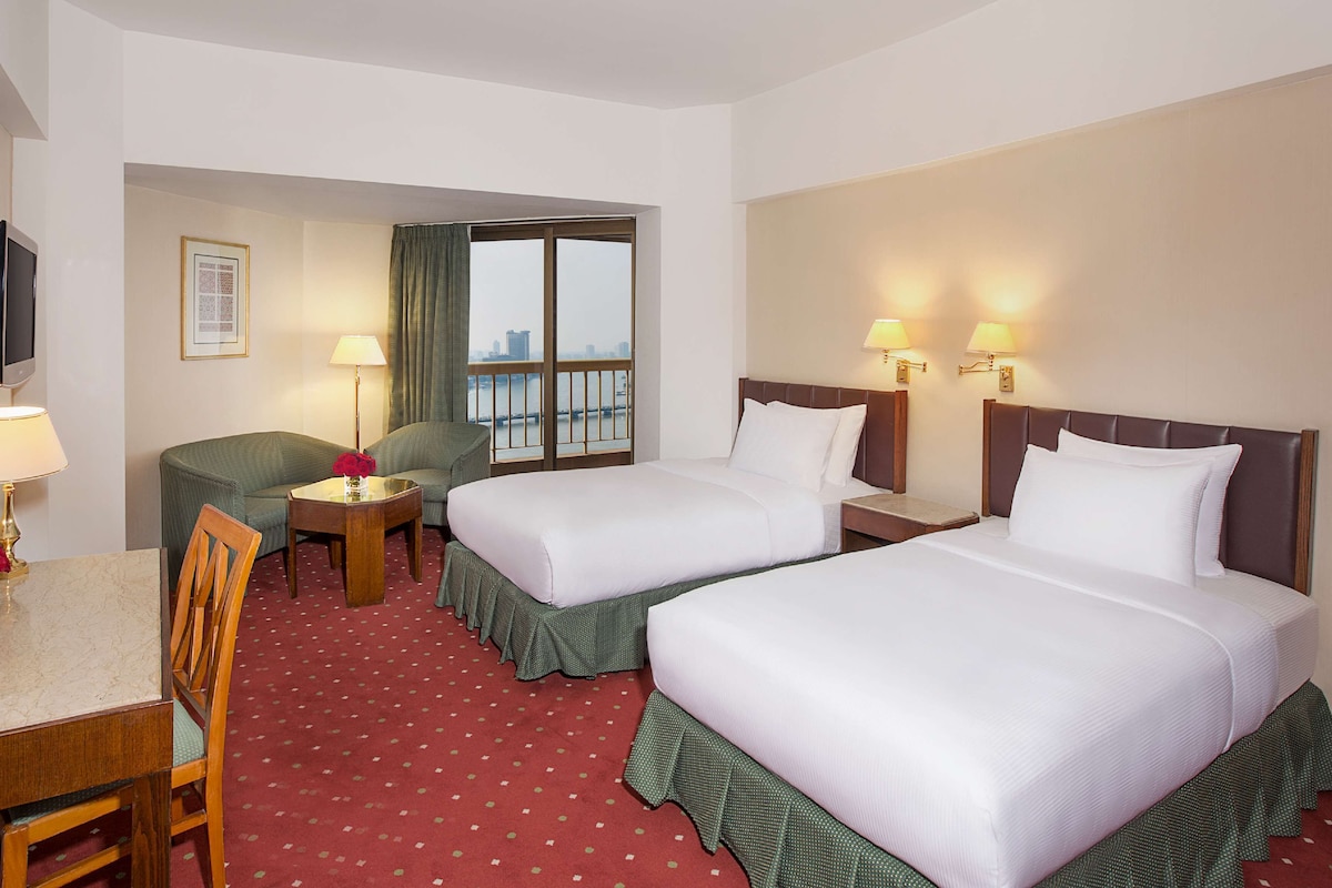 A hotel room with two beds, a desk, a chair, a TV, and a window with a view.