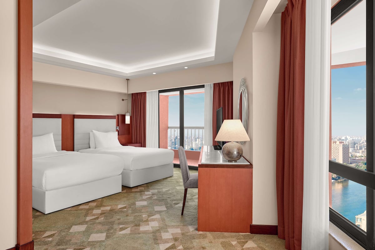 Une chambre d’hôtel avec deux lits, un bureau, une chaise et une grande fenêtre donnant sur la ville.
