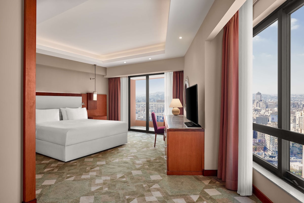 Une chambre d’hôtel avec un grand lit, un bureau avec une télévision, une chaise et une vue sur la ville grâce à de grandes fenêtres.