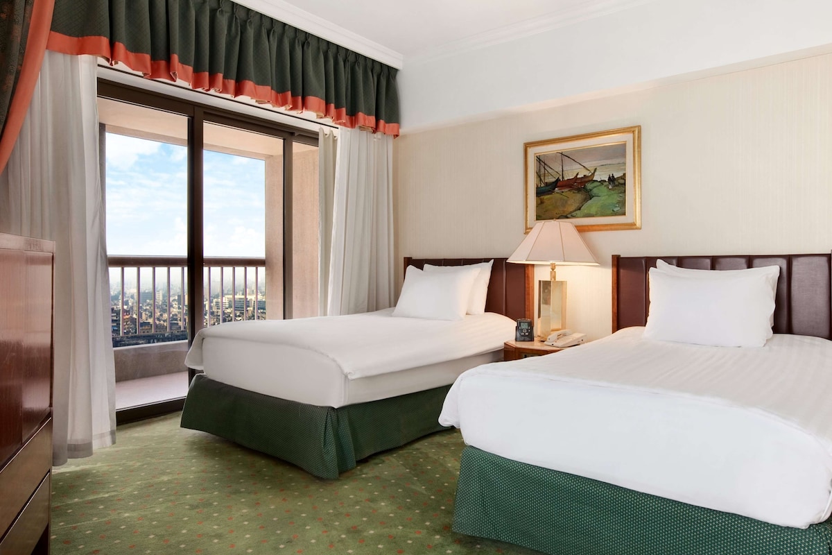 A hotel room with two beds, a large window with a view of the city, a painting on the wall, and a green carpet.
