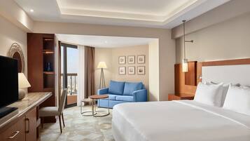 Quarto de hotel com uma cama grande, uma secretária com uma TV, um sofá azul, uma mesinha e vista para a cidade pela janela.