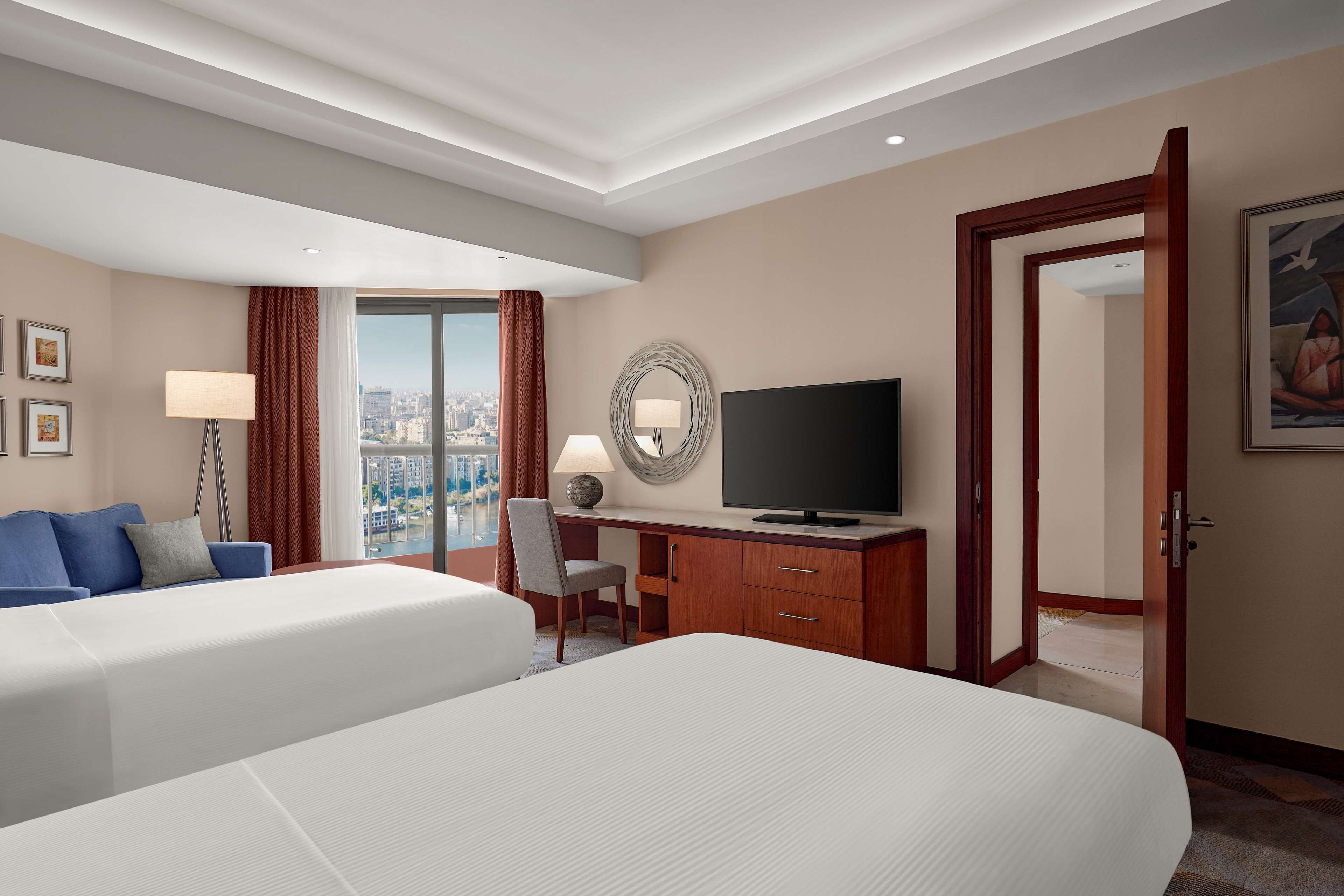 Habitación de hotel con dos camas, un escritorio con televisor, vistas a la ciudad y un espejo grande.