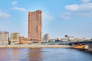 A tall hotel with a modern design, situated near a river with a bridge in the background.