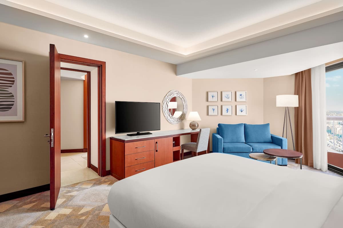 Une chambre d’hôtel avec un grand lit, un bureau avec une télévision, un canapé bleu, une petite table ronde et une vue sur la ville.