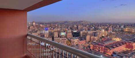 Un balcon avec vue sur la ville, offrant un panorama mêlant des immeubles de grande hauteur et une vaste structure illuminée au loin.