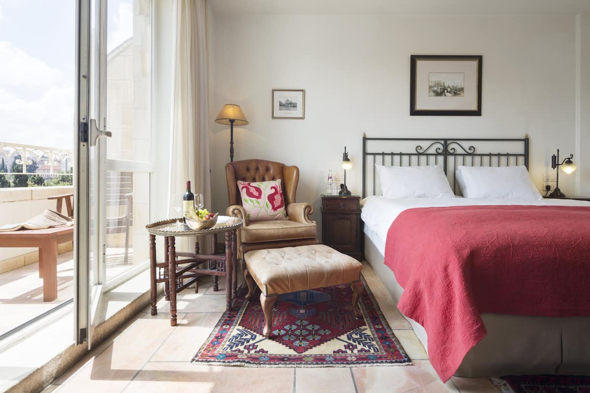 A hotel room with a bed, a chair, a table with a vase, and a view through a glass door.