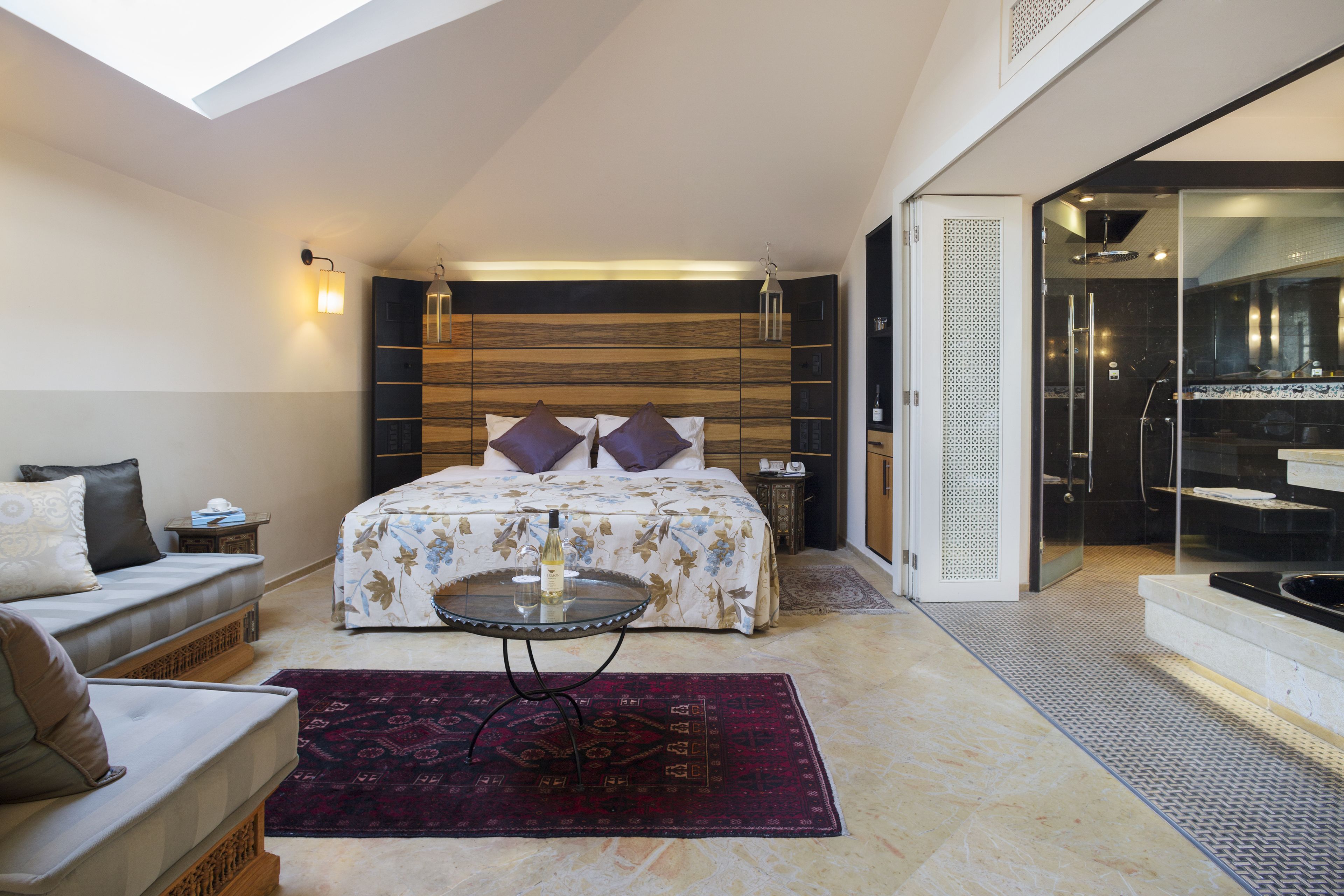 Ein modernes Schlafzimmer mit Dachfenster, einem großen Bett mit floralem Bettbezug, einer Duschkabine aus Glas und einem kleinen Tisch mit einer Flasche.