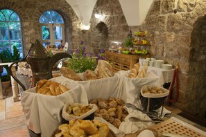 Ein Buffet mit verschiedenen Backwaren und einer Obstauslage in einem Raum mit Steinwänden und Rundbogenfenstern.