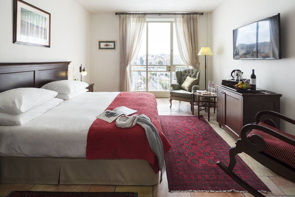 A hotel room with a large bed, a desk with a chair, a TV, and a view of the city through the window.