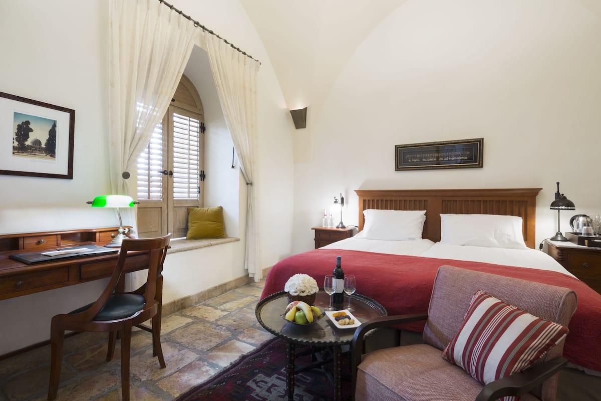 A hotel room with a bed, a desk, a chair, a small table with a bottle and glasses, and a window with curtains.