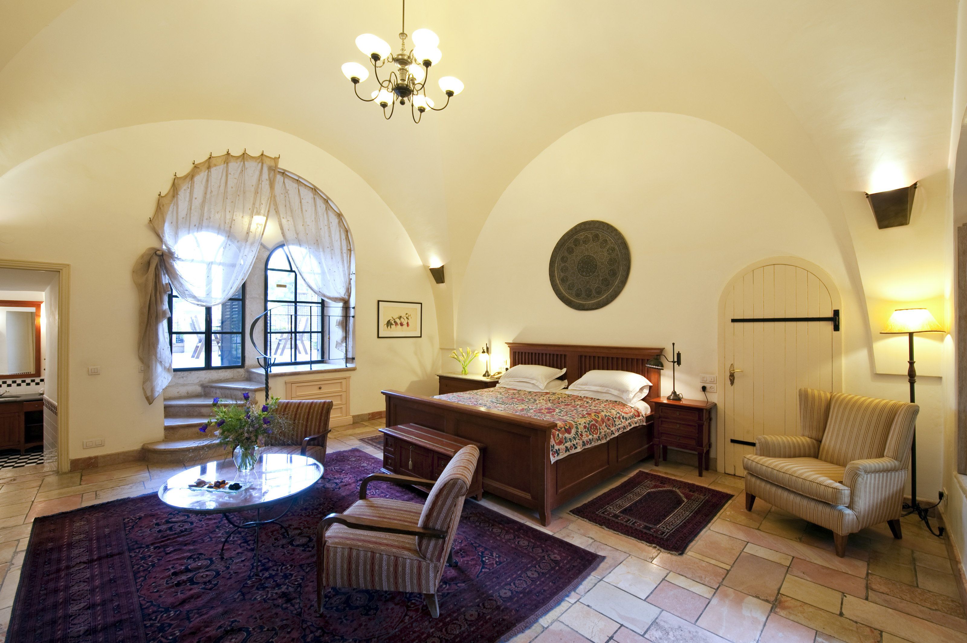 Una camera da letto con un letto grande, un angolo salotto con tavolo e sedie, un lampadario e vista all'esterno da una finestra.