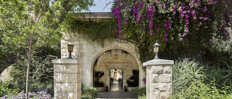 An arched stone entrance with a pathway leading through it, surrounded by lush greenery and flowering plants.